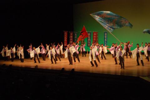 広いステージ上で、白と紫の衣装を着た数十人の人々が、赤い旗や巨大な青い旗を振りながら、和風の力強い踊りを披露している、賑やかな舞台発表の様子の写真