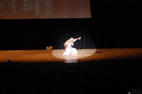 暗いステージの中央で、白い着物に長い髪の毛をなびかせた演者がスポットライトを浴びながら膝立ちになり、右腕を大きく広げるポーズを取っている、和風の舞踊か芝居のワンシーンを捉えた写真