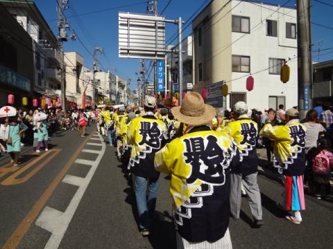 晴れた青空の下、大通りで行われている祭りの風景で、黄色と黒の法被を着た人たちが列をなして通りを進んでおり、沿道には提灯が飾られ多くの見物客が賑わっている様子を後方から捉えた写真