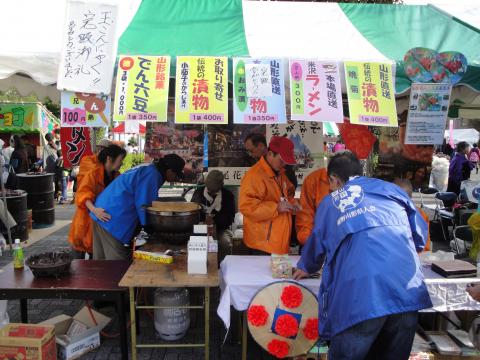 賑わうイベント会場で、緑と白のテントの下、オレンジ色と青色の服を着た人たちが「山形ラーメン」「山形名物」「お取寄せ品」などののぼりやメニューを掲げた屋台で調理や接客をしている様子を捉えた写真