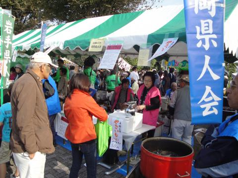 快晴の空の下、緑と白のストライプのテントの下で、ピンク色のベストを着た女性が来客に対応しており、手前には青いのぼり旗や赤い大きな鍋が見える、地方の物産展のようなイベント風景の写真