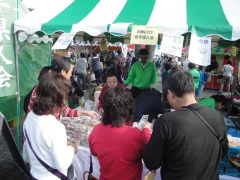 多くの人で賑わう屋外イベント会場で、緑と白のテントの下に設けられたブースにおいて、客と売り手が和菓子のようなものを販売するテーブルを囲んで活発にやり取りしている様子の写真