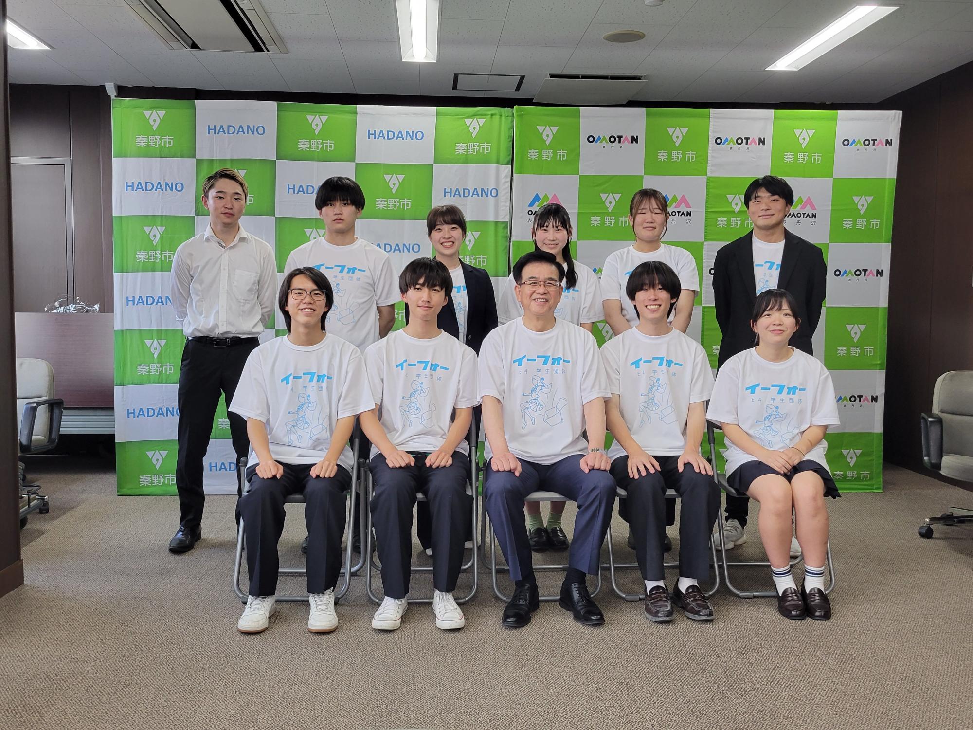 緑と白のチェック柄に「秦野市」のロゴが並ぶ背景の会議室で、「イーフォー」と書かれた白いTシャツや白いシャツを着た学生たちと、中央の椅子に座る市長が、カメラに向かって並んで写っている集合写真
