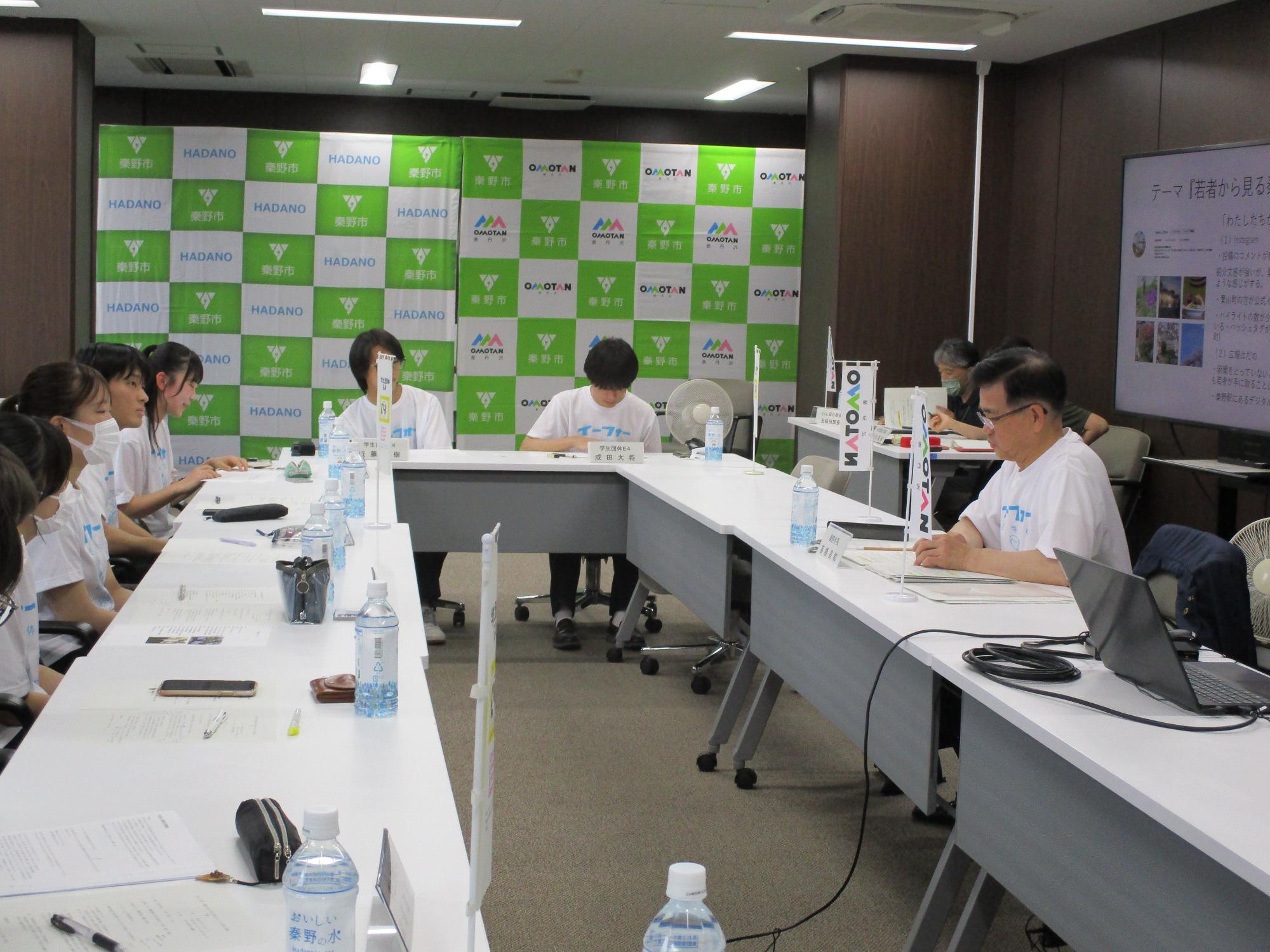 緑と白のチェック柄に「秦野市」のロゴが並ぶ背景の会議室で、コの字形に配置された白いテーブルを、白いTシャツを着た生徒や大人たちが囲み、右側の席に座る市長が資料に書き込みながら、議論に耳を傾けている様子の写真