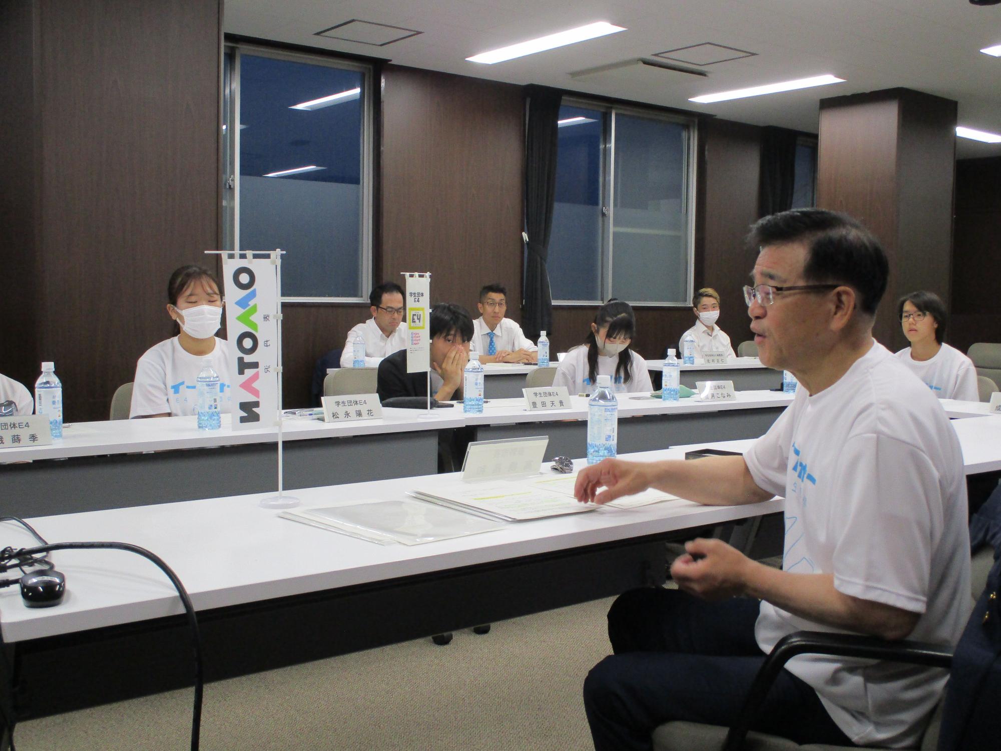 木目調の壁の会議室で、白いTシャツを着た市長が手前のテーブルに座って身振り手振りを交えながら話しており、その奥のテーブルにはマスク姿の生徒数名とポロシャツ姿の大人たちが座って真剣に耳を傾けている意見交換会の風景写真