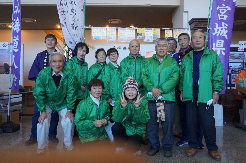 屋内イベント会場らしき場所で、緑色のジャンパーを着た11人の中高年男女のグループがカメラに向かって並んで立っており、中央の女性2人がかがんでピースサインをしていて、背景には「宮城県」などの文字が入ったのぼりやポスターが並ぶ集合写真