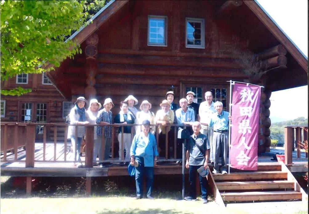 青空の下、多数の高齢者が、ログハウス風の木造建物の広いウッドデッキに集まり、赤い縦幕に「秋田県人会」と書かれたのぼりの横で、穏やかな表情を浮かべている屋外での集合写真