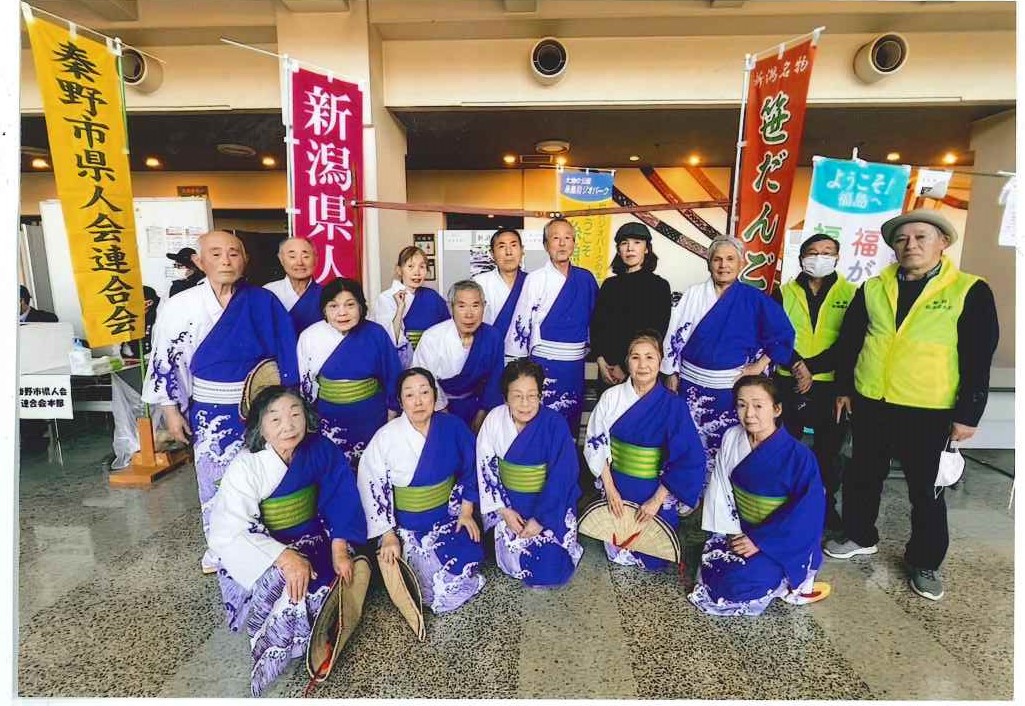 「新潟県人会」や「ようこそ福島へ」などと書かれたのぼりが立つ屋内イベントスペースで、浴衣のような和装の衣装を着た13人の高齢者が、男性数名と女性のグループで並び、うち女性7人がしゃがんで扇子のようなものを持っている集合写真
