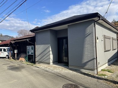 細い生活道路沿いに建つ、濃いグレーの外壁と黒い屋根が特徴のモダンな平屋の建物が写る写真