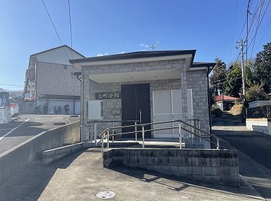 青空の下、ライトグレーの石目調の外壁を持つ建物の正面に「上町会館」と書かれた看板があり、その手前には段差をなくしたスロープと手すりが設置された玄関アプローチが広がる写真