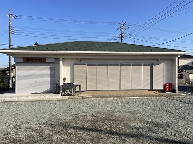 明るい日差しの下、白い壁と濃い緑色の傾斜屋根を持つ平屋の建物の正面に、「飯坂町役場」と書かれたシャッター付きの小窓と、大きなパネル式のシャッターが並び、手前には砂利敷きの広い駐車場が広がる写真