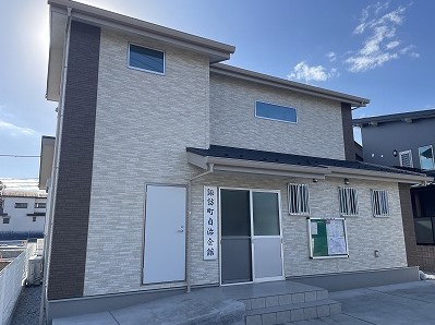 淡いベージュと濃い茶色のツートンカラーの外壁を持つ、玄関ポーチと二階建ての部分が組み合わされたモダンな集合住宅のような建物が、正面のガラス戸の上に「諏訪町自治会館」と書かれたプレートを掲げている写真