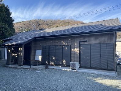 穏やかな日差しの下、濃い茶色の壁と黒い屋根を持つモダンな平屋の建物が、広々とした砂利敷きの駐車場に面して建ち、窓には濃い色のルーバー状の目隠しが取り付けられている写真