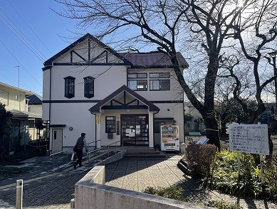 白壁と濃い木目調の装飾を持つ二階建ての建物が、日差しを浴びて建っており、手前には自動販売機や掲示板が設置され、左側には人物が歩くスロープ付きの通路が設けられている写真