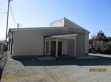 淡いベージュの壁と黒い屋根を持つ、手前に屋根付きのポーチと二枚の暗色のドアを備えた平屋建てのモダンな建物が、広大な砂利敷きの駐車場に面して建ち、青空の下で明るい日差しを浴びている写真