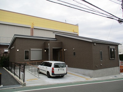 黒茶色のサイディングの外壁と黒い傾斜屋根を持つモダンな平屋の建物が、白いワゴン車一台分の駐車スペースとスロープを備えている建物の写真