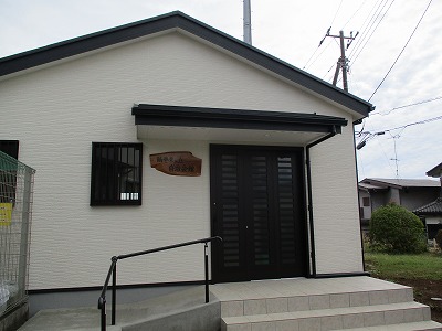 シンプルな切妻屋根とアイボリーのサイディング外壁が特徴の平屋の建物が、正面の黒い両開きドアの上に「鶴巻東ヶ丘自治会館」と書かれた木製の看板を掲げており、玄関前には階段と手すりが設置されている写真