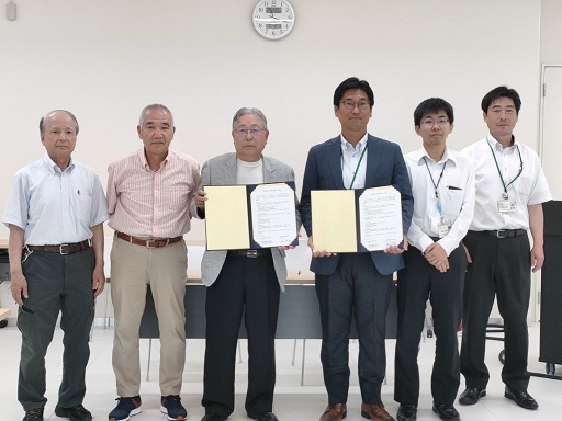 白い壁と時計の下で、6人の男性が並び立ち、中央の二人がそれぞれ白い紙の入った薄黄色のファイルや書類を持って正面を向いている調印式または協定締結の様子の写真