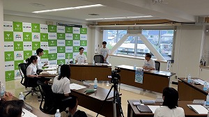 窓のある明るい会議室で、緑色のロゴが並んだ背景の前に複数の人が向かい合って座る大きなテーブルがあり、白いポロシャツを着た男性が生徒らしき人々に向けて立って話している様子の写真