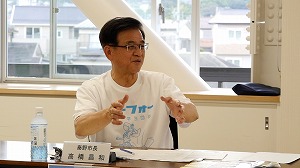 窓から外の景色が見える明るい会議室で、「イーフォー」と書かれた白いTシャツを着た市長が、手を動かしながら熱心に話している様子の写真