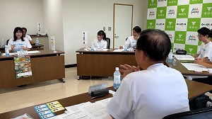 緑と白のロゴマークが並ぶ背景の会議室で、白いTシャツを着た数名の生徒や関係者らしき人々がテーブルを囲み、手前で後姿の市長が相槌を打つ中、向かい側の女性が笑顔で話している様子を捉えた写真