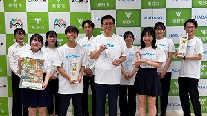 緑と白のチェック柄に「OMOTAN 栗野市」のロゴが並ぶ背景の前に、「イーフォー」と書かれた白いTシャツを着た市長を中央に、ポロシャツ姿や白いTシャツ姿の男女合わせて10人の生徒や関係者が並び、それぞれポスターや箱のようなものを手に持って笑顔で写る集合写真