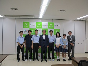 緑と白のロゴが並んだ背景の会議室で、スーツ姿の市長を中央に、ポロシャツや制服姿の男子生徒4人と女子生徒2人、スーツ姿の別の男性1人の計8人が、カメラに向かってまっすぐ立って微笑んでいる集合写真