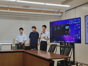 会議室で、茶色い机の前に立つ男子生徒3人が、左端の生徒はポロシャツ、中央と右端の生徒は白いワイシャツ姿で、右端の生徒はマイクを持って発言しており、パソコンのデスクトップ画面が大きなモニターに映し出されている発表風景の写真