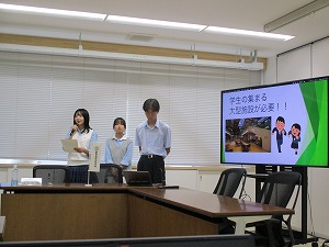 会議室の茶色い机の前に、制服姿の女子生徒2人と白いワイシャツ姿の男子生徒1人が立ち、右側のモニターに「学生の集まる大型施設が必要!!」と書かれたスライドを映しながら、女子生徒がマイクを持って発表している様子の写真