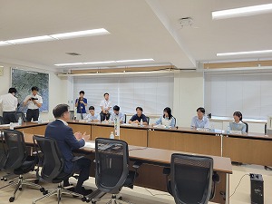 白い壁の会議室で、スーツ姿の市長が背を向けて手前の席に座り、その奥の長いテーブルを挟んで、ポロシャツ姿の生徒や大人たちが着席し、さらに奥では数名の報道関係者らしき人々が撮影や取材をしている様子の写真