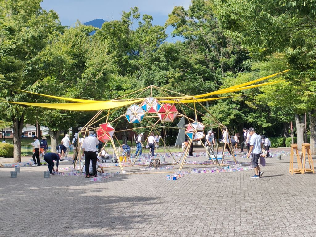 夏の日差しの下、公園の木々に囲まれた広場で、大勢の人々が竹とロープで作られたドーム型の骨組みに赤と青の星型の飾りや黄色の布を取り付けている、祭りやイベントの準備風景を捉えた写真