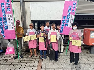 屋外の壁の前で、ピンク色のエプロンを着用した男女7人が、募金箱や感謝状のような書類を手に持ち、「はだの市民活動団体連絡協議会」と書かれたピンクの旗を掲げて立っている様子の集合写真