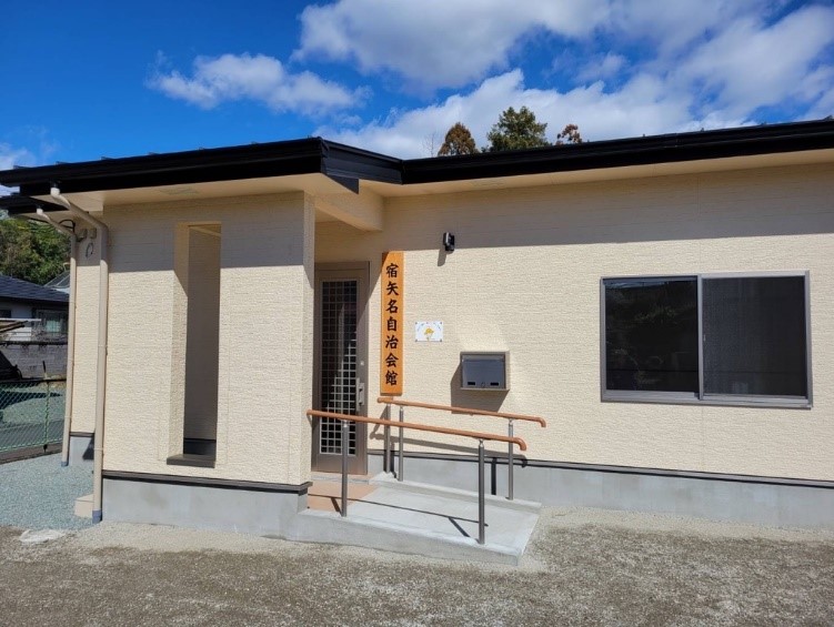 青空の下、明るいベージュの外壁と黒い屋根を持つ平屋の建物が、玄関ポーチに縦書きで「宿矢名自治会館」と書かれた木製の看板を掲げ、手すり付きのスロープが設けられたバリアフリー対応の玄関が特徴的な写真