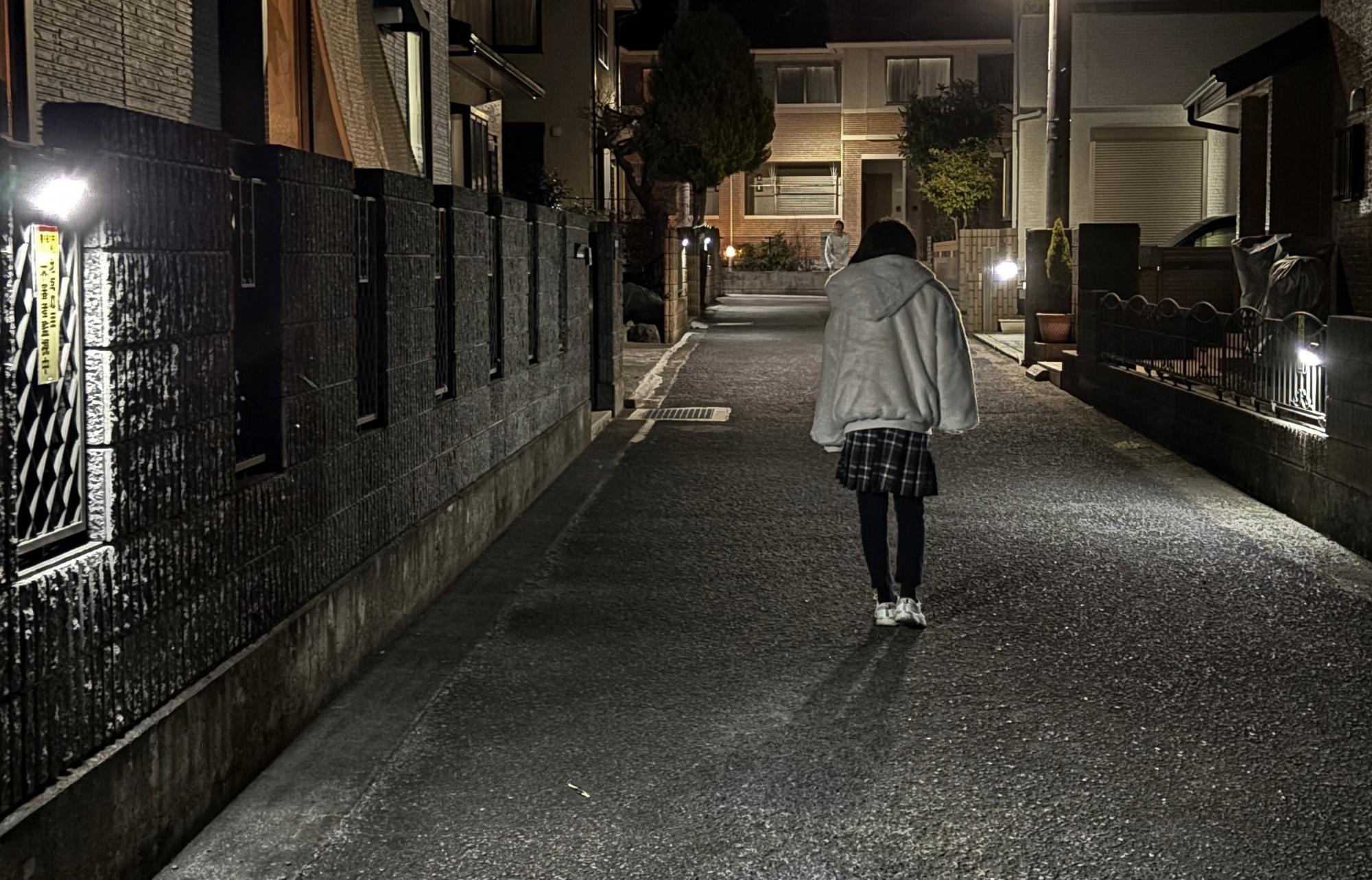 夜間に住宅街の細い舗装された道を歩く若い女性を後ろから捉えた写真で、左右の塀には防犯灯のような照明が取り付けられており、道の奥の建物も街灯に照らされている写真