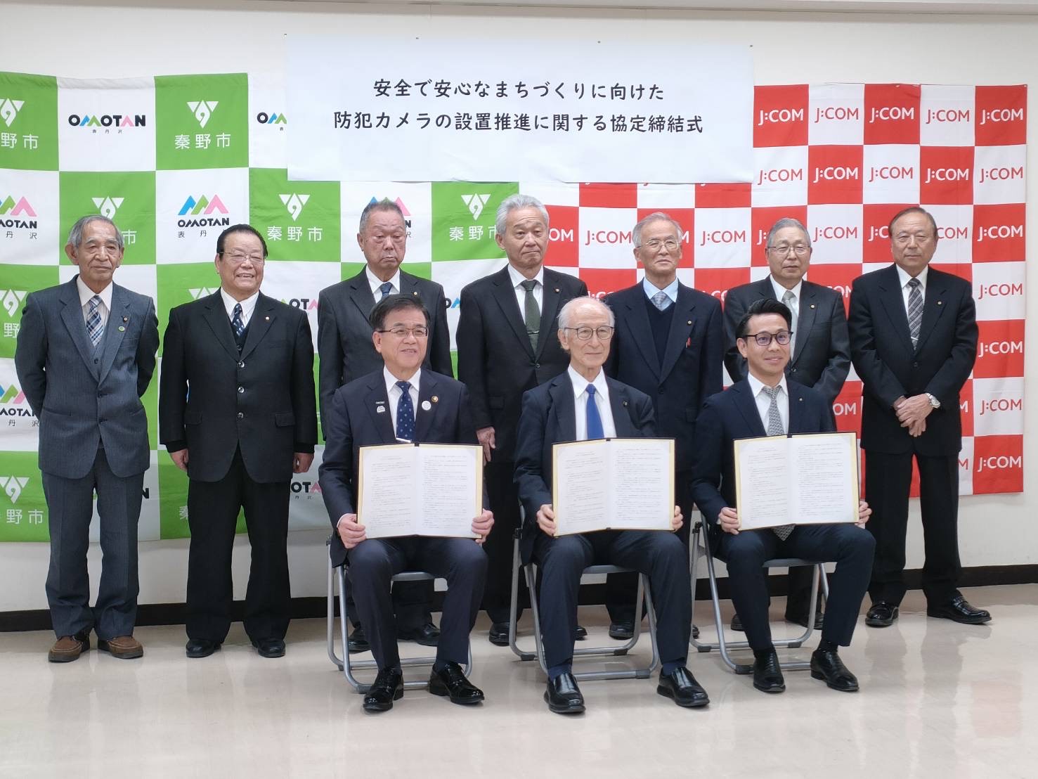 「安全で安心なまちづくりに向けた防犯カメラの設置推進に関する協定締結」と書かれた横断幕の下で、スーツ姿の複数の男性たちが、座って協定書を開いて正面に見せている写真