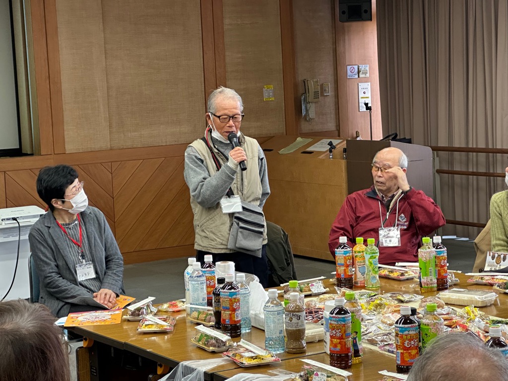 中央に立つ白いベスト姿の高齢の男性がマイクで発言しており、その両隣にはテーブルに並べられたペットボトルや弁当の前で話を聞く男女が座っている様子の屋内写真
