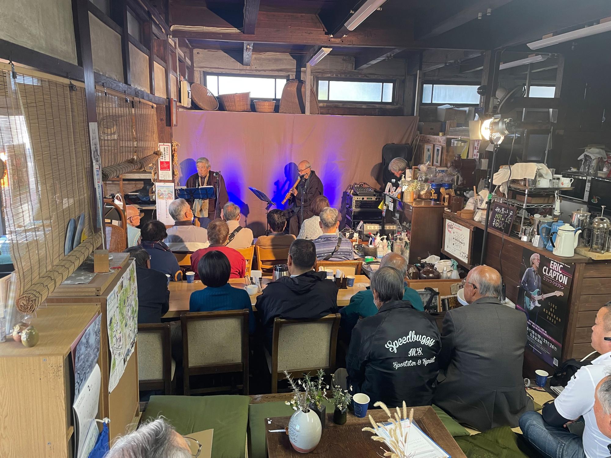 背景の紫色の照明に照らされたステージの上で、2人のミュージシャンが、集まった聴衆に向かって、尺八を披露しているライブ会場の写真