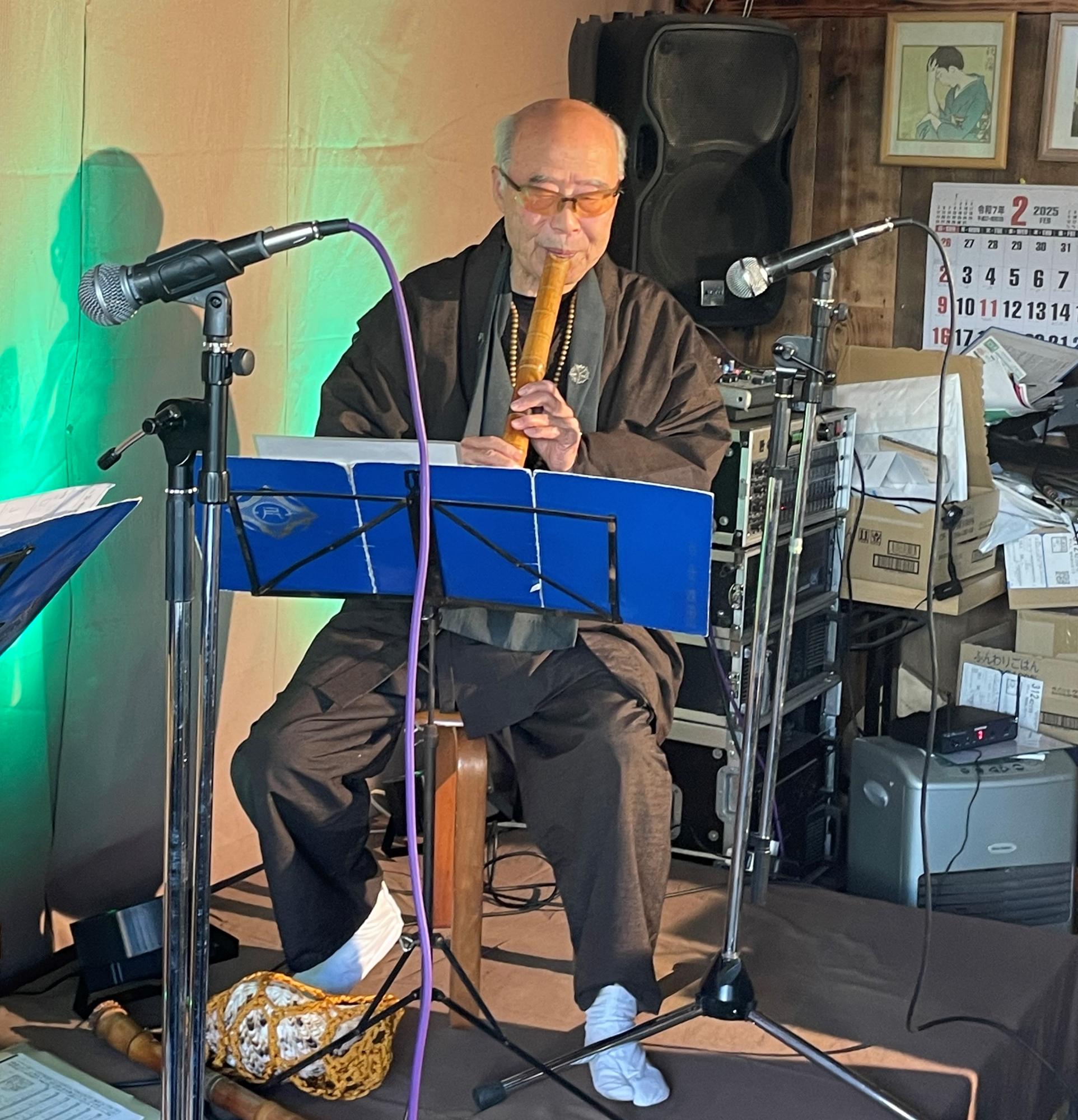 黒い着物にサングラスをかけた星秦山さんが、照明で緑色に照らされた背後の機材の前で、譜面台に置かれた楽譜を見ながら尺八を演奏しているライブパフォーマンスの写真