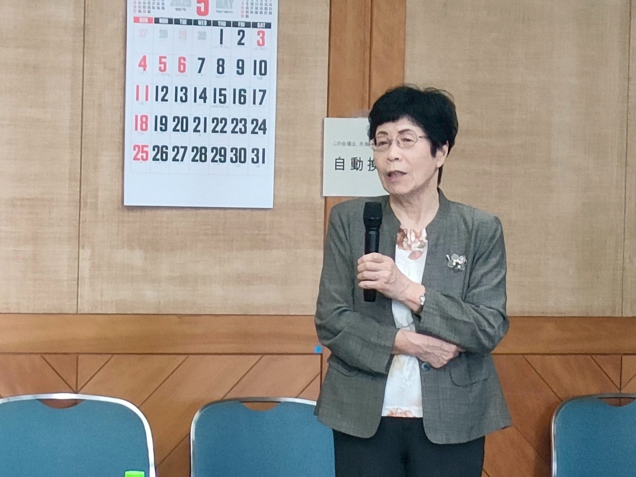 グレーのジャケットと花柄のブラウスを着用した女性が、腕を組みながらマイクを持って話している様子を捉えた写真