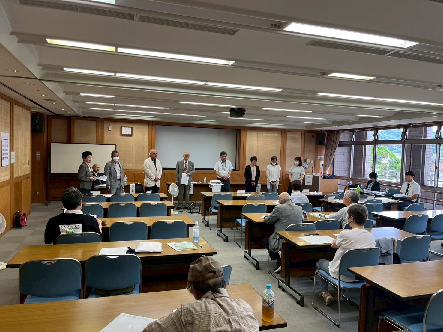 会議室で、大勢の参加者が机と椅子に座って聴講する中、白いスクリーン前の壇上に複数の発表者と関係者が並び挨拶をしている様子の写真