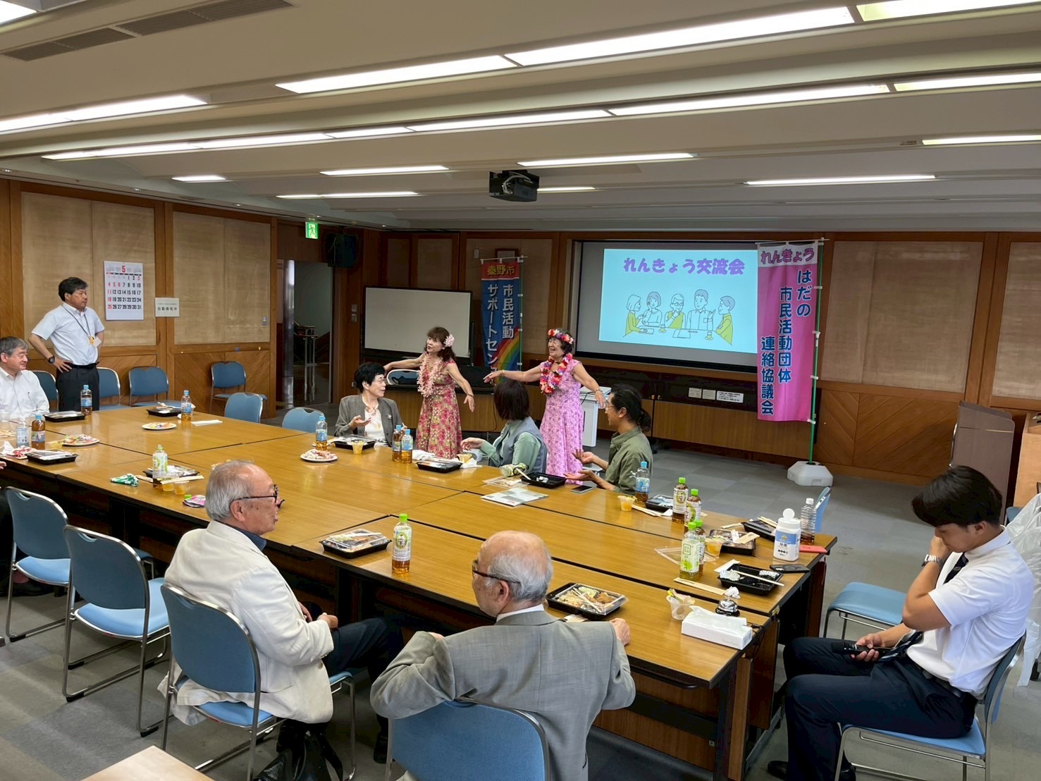 広々とした会議室で、長机を囲んで座る複数の参加者の前で、花飾りとフラダンス風の衣装を身に着けた二人の女性が踊りを披露しており、壁のスクリーンには「れんきょう交流会」と表示されている交流会の写真