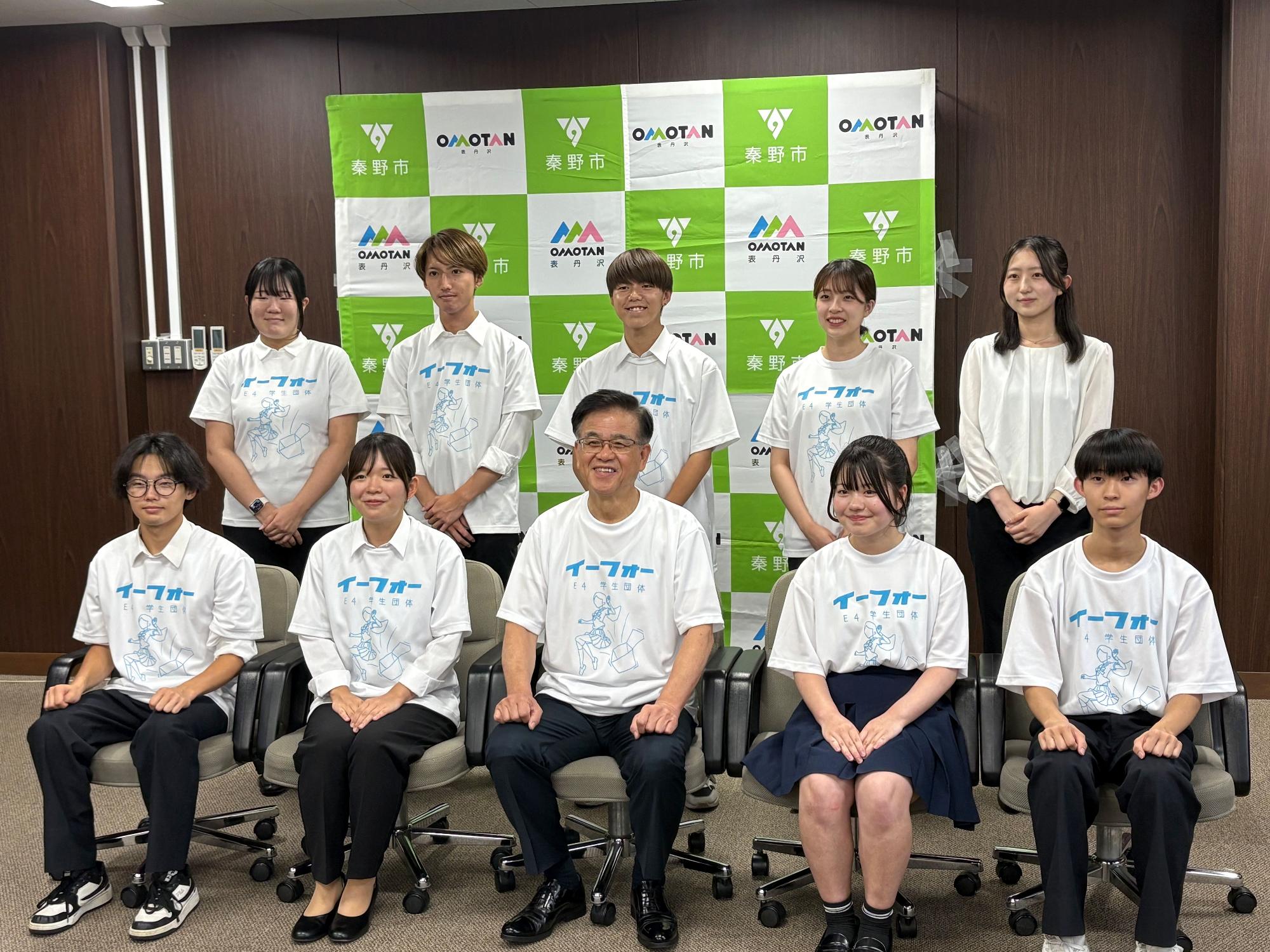 「イーフォー」と書かれたTシャツを着た生徒9人と、中央に座るスーツ姿の男性1人が、緑と白のチェック柄に「OMOTAN 栗野市」と書かれたロゴマークの背景の前に並んで、カメラに向かって微笑んでいる集合写真