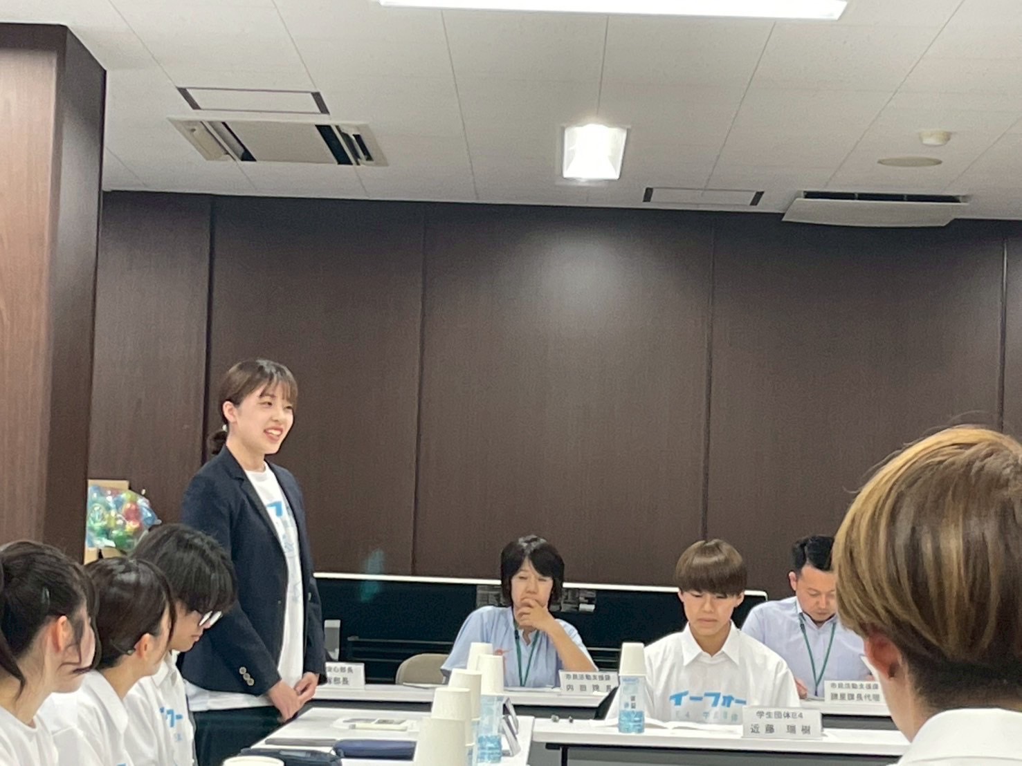 薄茶色の木目調の壁の会議室で、白いTシャツを着た数名の生徒や、黒いジャケットの女性、ポロシャツ姿の大人たちがテーブルを囲んで座る中、左端の女性が立ち上がって笑顔で発言している会議風景の写真