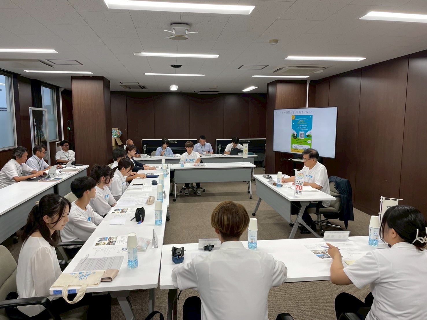 木目調の壁の会議室で、白いTシャツを着た数名の生徒たちがコの字形に配置された机に座り、プロジェクターに映された資料を見ながら、向かい側のポロシャツ姿の市長と真剣に意見交換をしている会議の風景写真