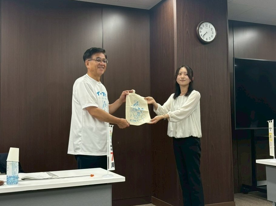 木目調の壁の会議室で、白いTシャツを着た市長と白いブラウスを着た若い女性が、にこやかに向かい合って、キャラクターがプリントされた布製の袋のようなものを両手で受け渡ししている様子の写真