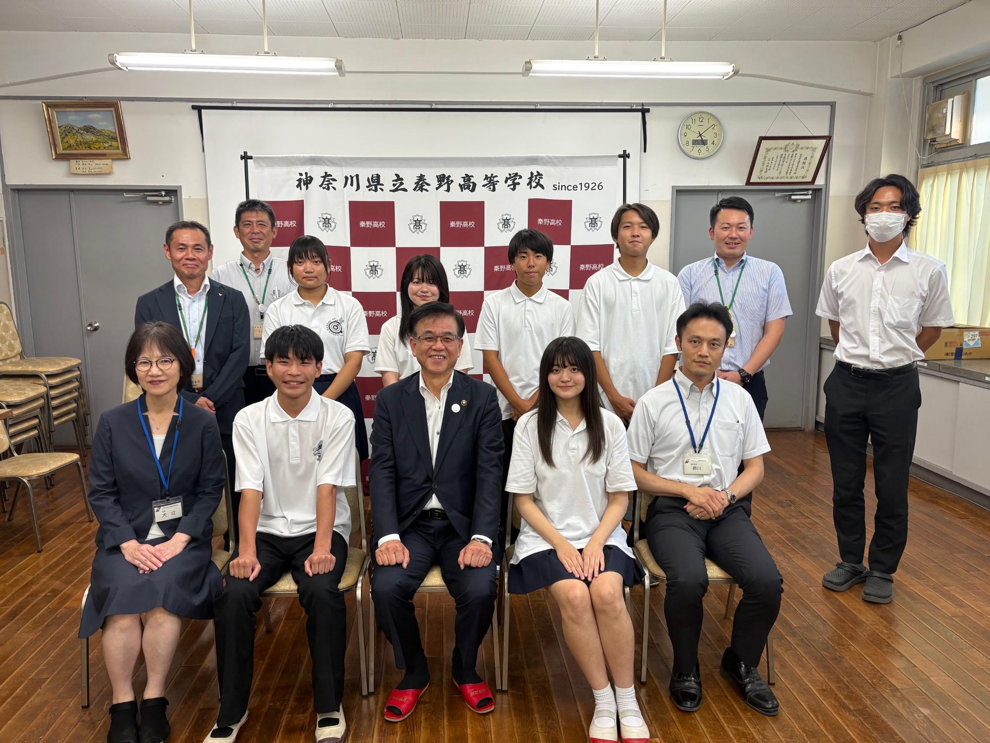 神奈川県立秦野高校の校章を背景に、スーツ姿の教員らしき大人7人と、白いポロシャツと紺のスカートまたはズボン姿の学生が、椅子に座る者と立つ者に分かれて並び、カメラに向かって笑顔で写る集合写真