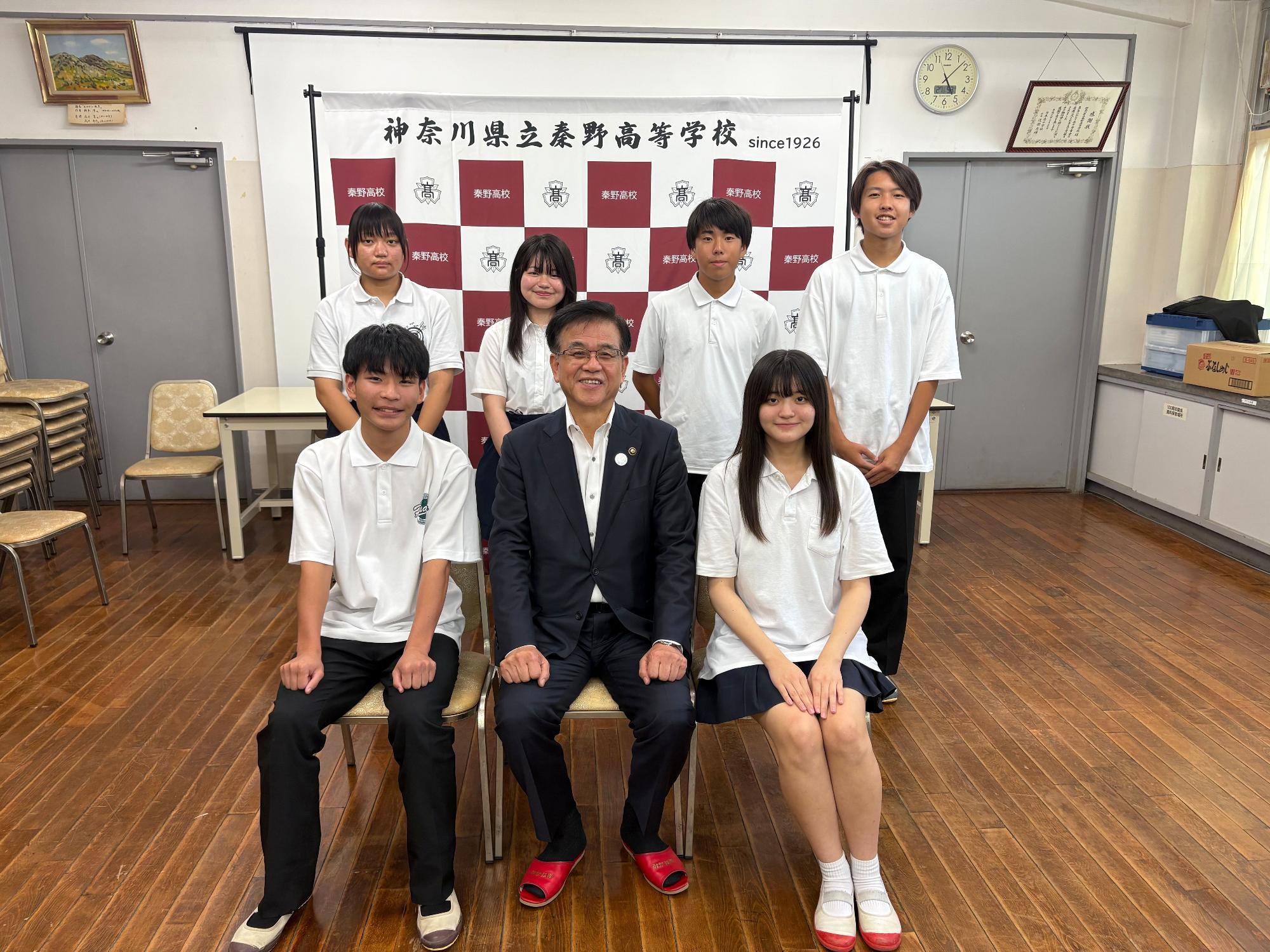 神奈川県立秦野高校の校章を背景に、スーツ姿の市長が中央の椅子に座り、白いポロシャツと紺のスカートまたはズボンの制服を着た男子生徒3人と女子生徒3人が、その周囲に立ったり座ったりして笑顔で並んでいる集合写真