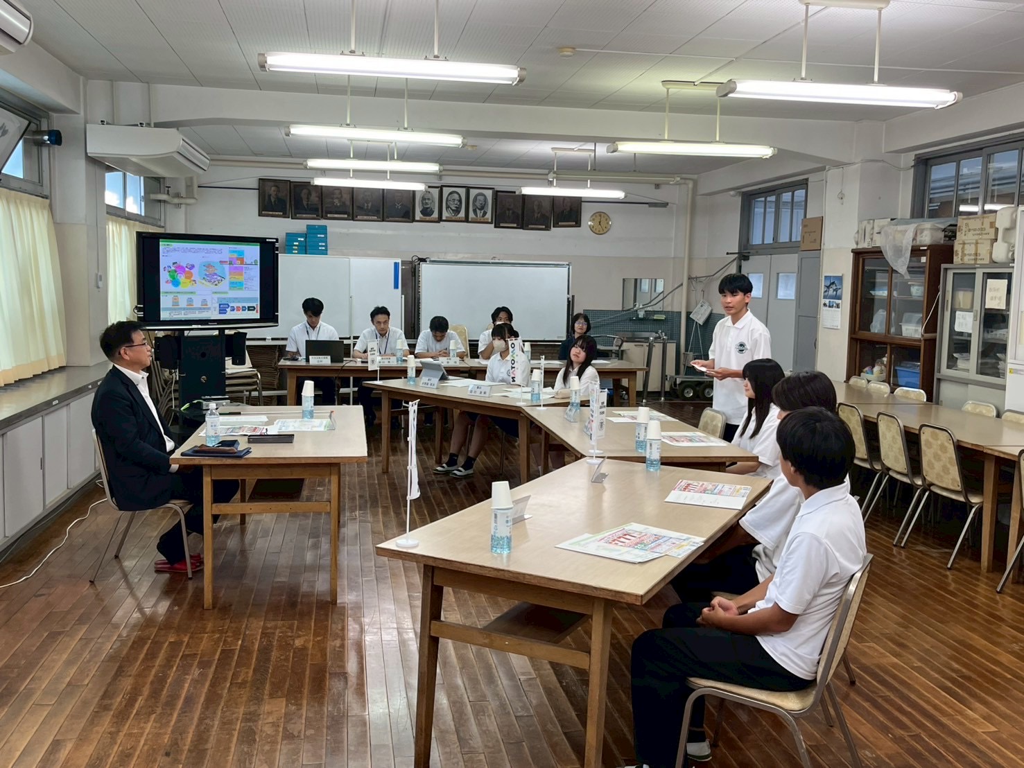 広くて明るい木目調の床の教室で、スーツ姿の市長と白いポロシャツの生徒たちが配置された机を囲み、1人の男子生徒が立って発表している様子の写真