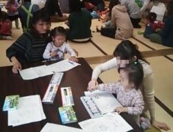 畳敷きの広間で、二組の母子がテーブルを囲んで座り、筆記用具を使い大人が子供の手に触れて一緒に何かを書こうとしている様子の写真