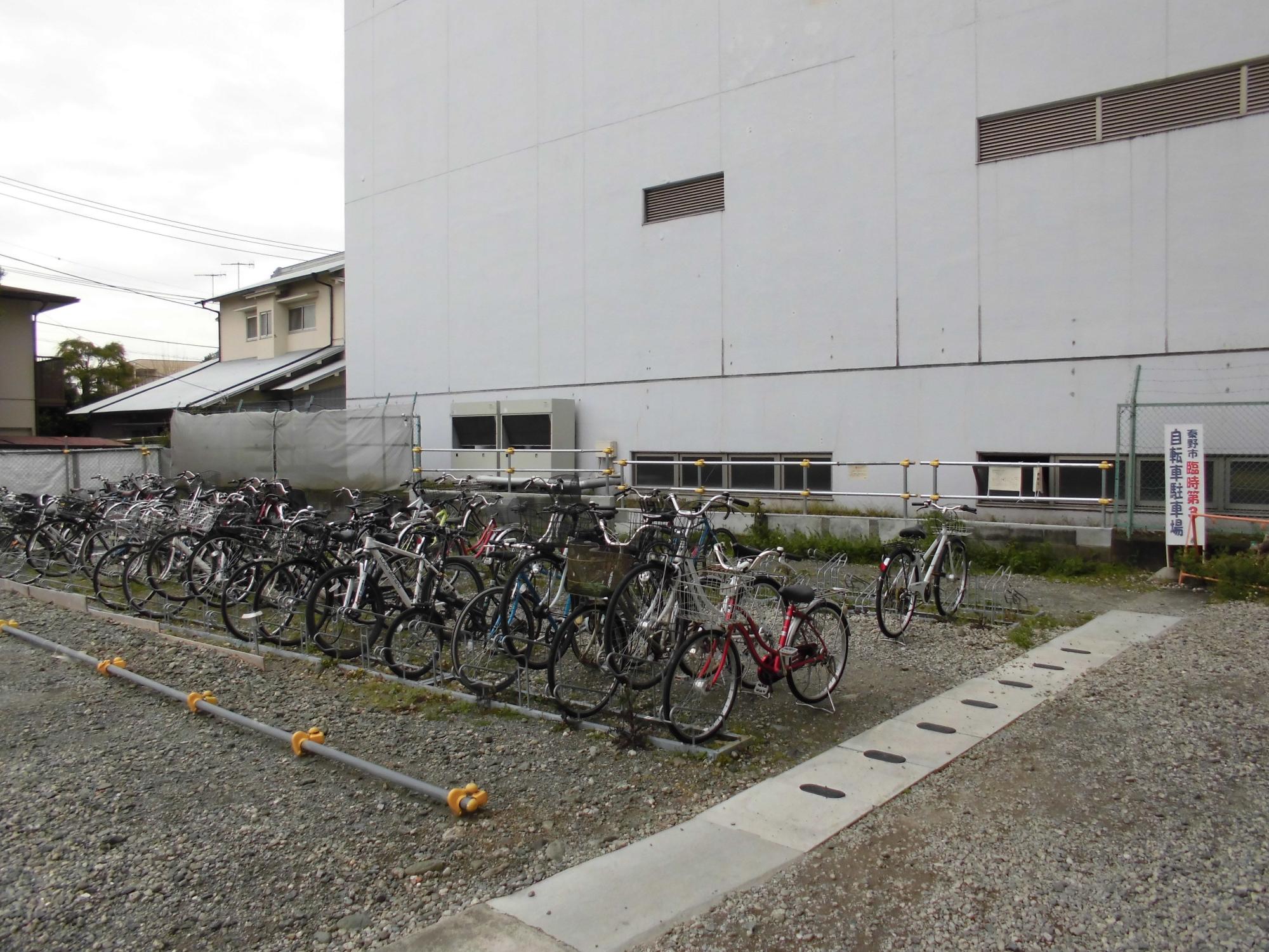 大きな白い壁の建物の横の砂利敷きの敷地に、多数の自転車が駐輪ラックに密集して停められており、手前にはコンクリートの排水溝と黄色のパイプで仕切られたラインが見える屋外駐輪場の写真
